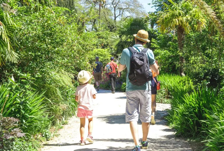 This image depicts a man with his young daughter walking down a path and towards a botanical garden. They're both dressed in summery clothes with sun hats on.