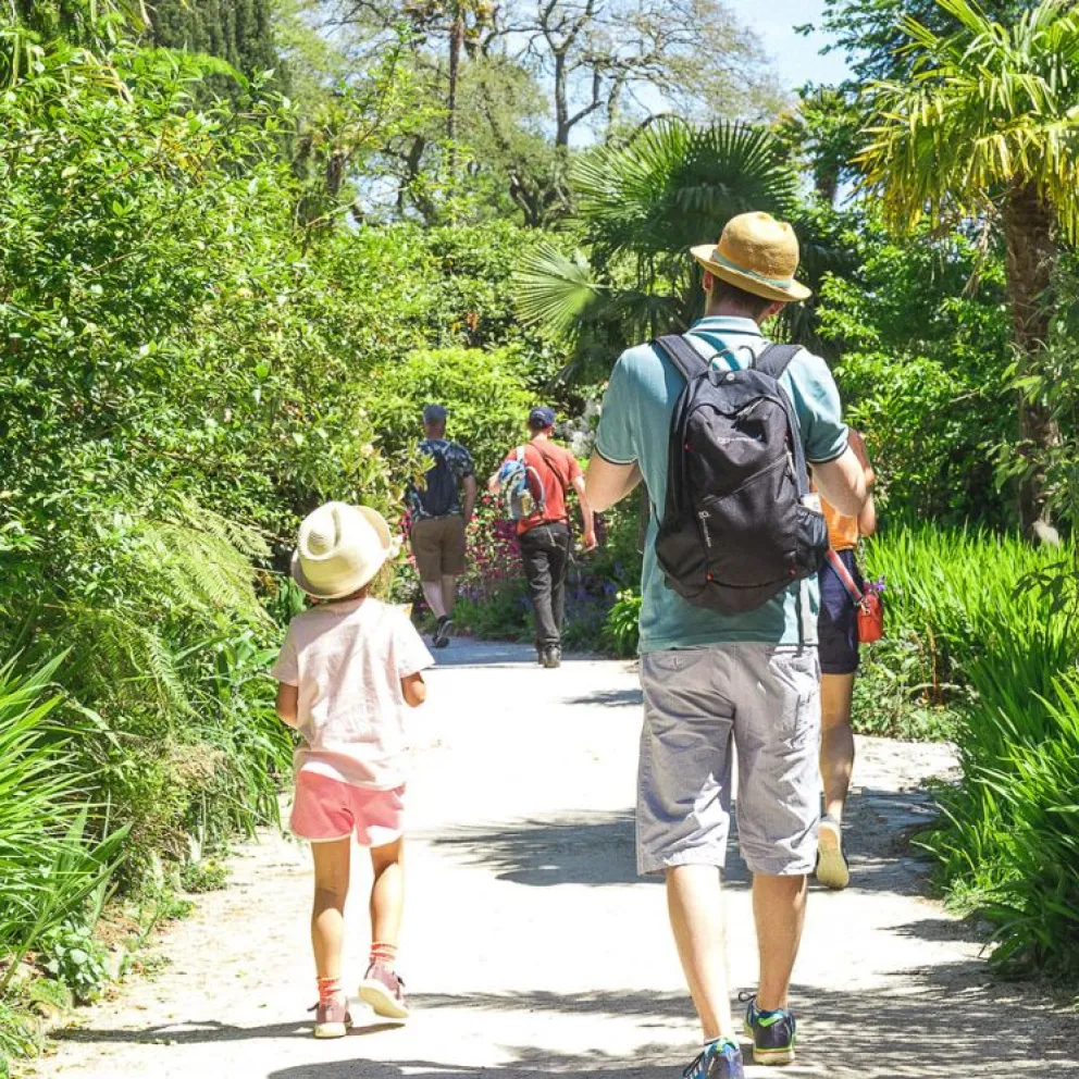 This image depicts a man with his young daughter walking down a path and towards a botanical garden. They're both dressed in summery clothes with sun hats on.