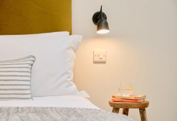Close up of bed with white bedding and a small wooden stool as a bedside table with stacked books and a glass of water.