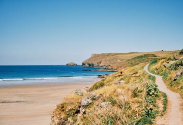 Views over Pentire Point from Polzeath, with golden sands and footpath in the foreground and sparkling ocean and clifftops in the background.