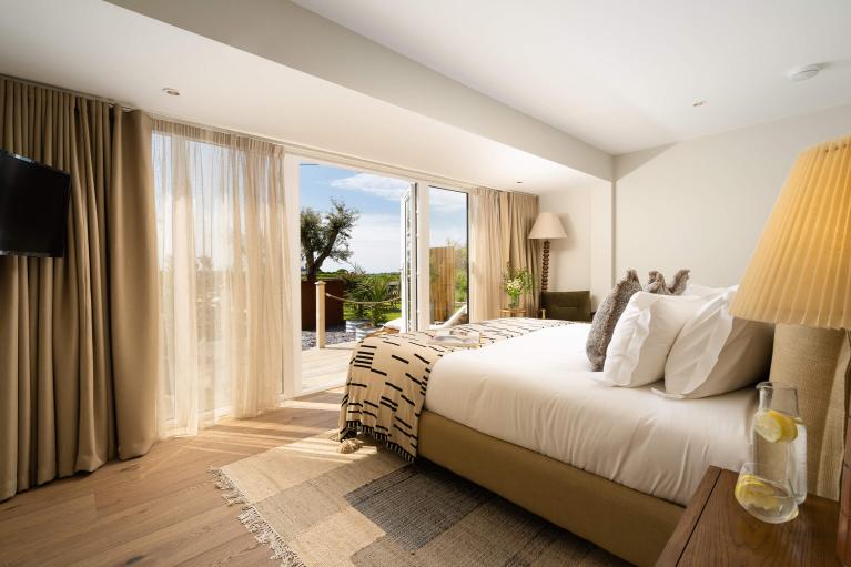 Bedroom with bed made with white linen sheets, a cream bedspread and brown linen cushions. The interiors are neutral and there's French doors opening up onto a decked terrace.