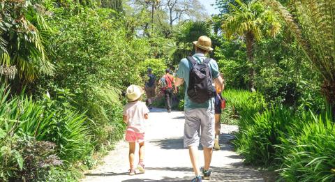 This image depicts a man with his young daughter walking down a path and towards a botanical garden. They're both dressed in summery clothes with sun hats on.