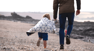 A father and son holding hands on the beach, walking away from the camera