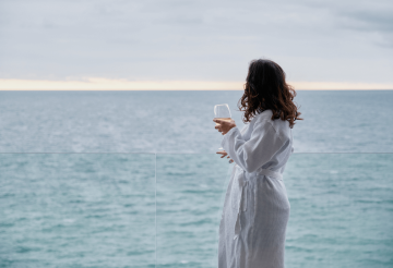 A woman in a white robe, holding a glass of wine and looking out to sea